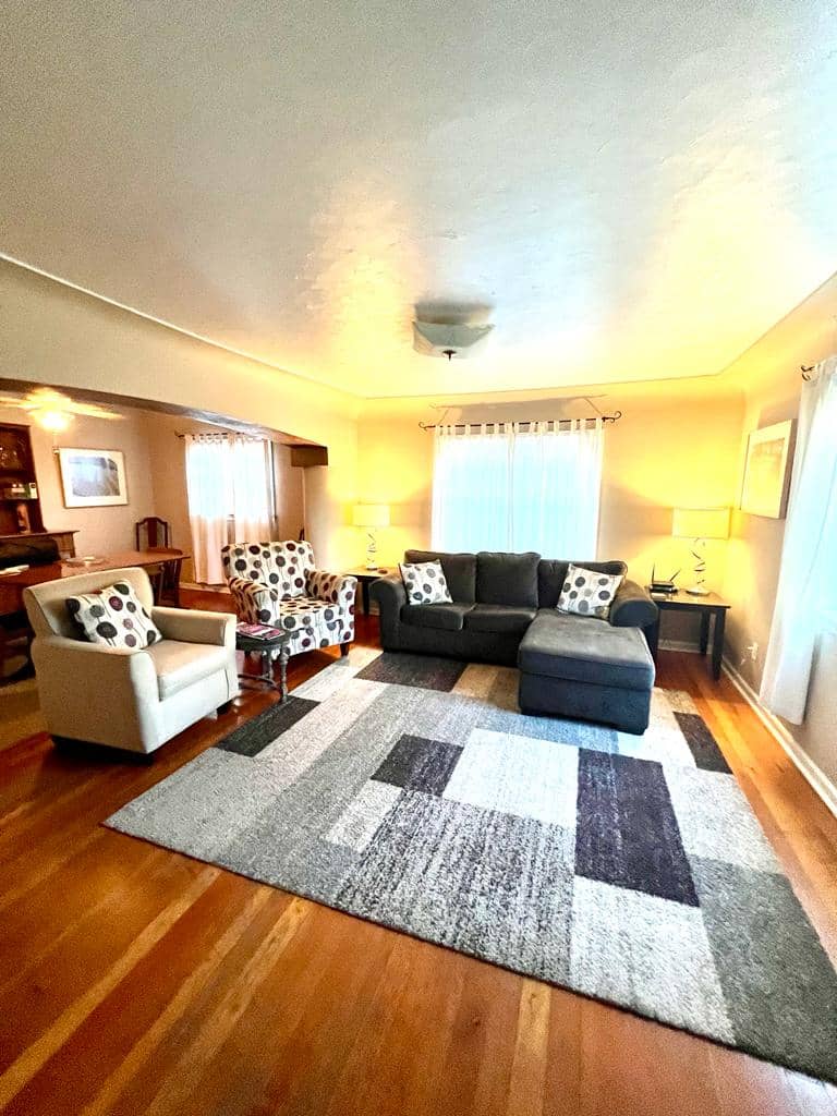 Living room with hardwood flooring, couch, two cloth chairs, television, and a big rug